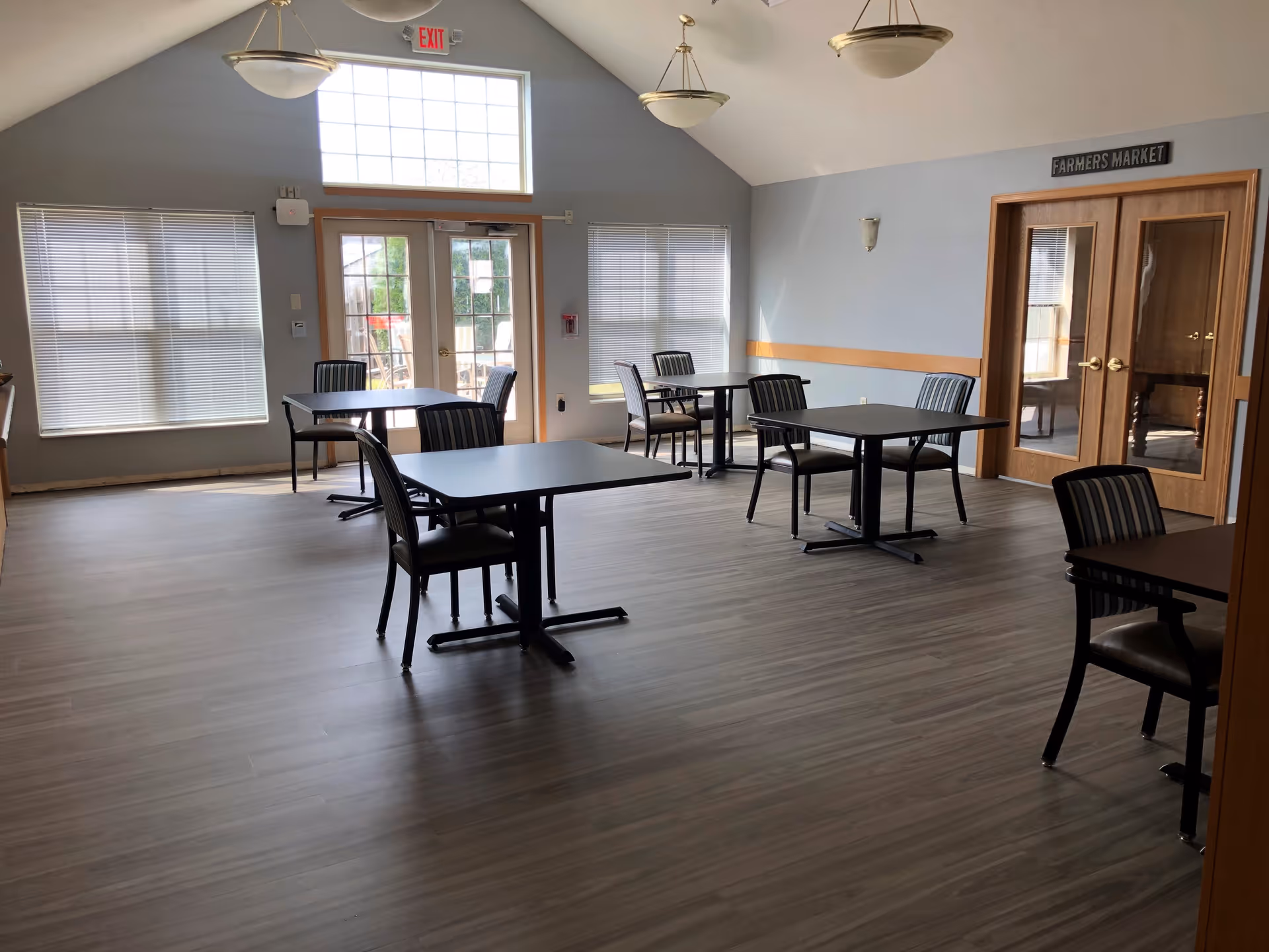 A bright dining room with several square tables and chairs spaced apart on a wood floor. The room has large windows with blinds, a set of double glass doors with an exit sign above, and a sign above another set of double wooden doors that reads 'Farmers Market'. The walls are light gray with a wooden chair rail, and there are hanging light fixtures from the vaulted ceiling.