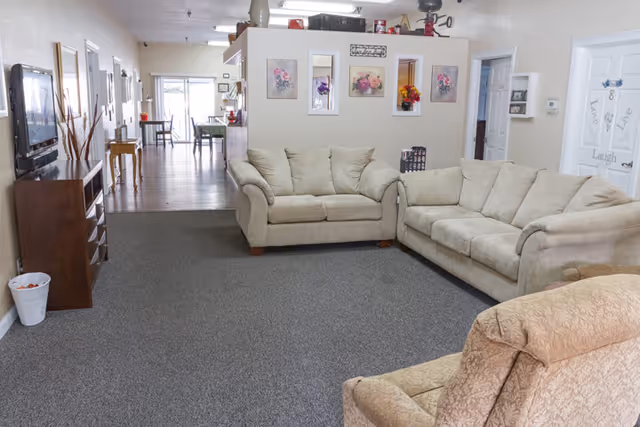 Communal living room with beige sofas and an armchair facing a wall-mounted TV, decorative wall art on a partition, and a dining area visible in the background.