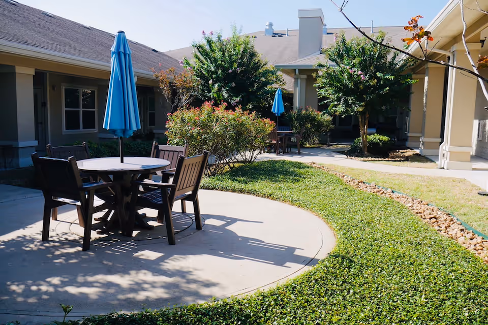 Outdoor courtyard area at Orchard Park at Victory Lakes Assisted & Senior Living featuring round tables with blue umbrellas and wooden chairs, surrounded by green shrubs, trees, and a building with beige walls and windows in the background.