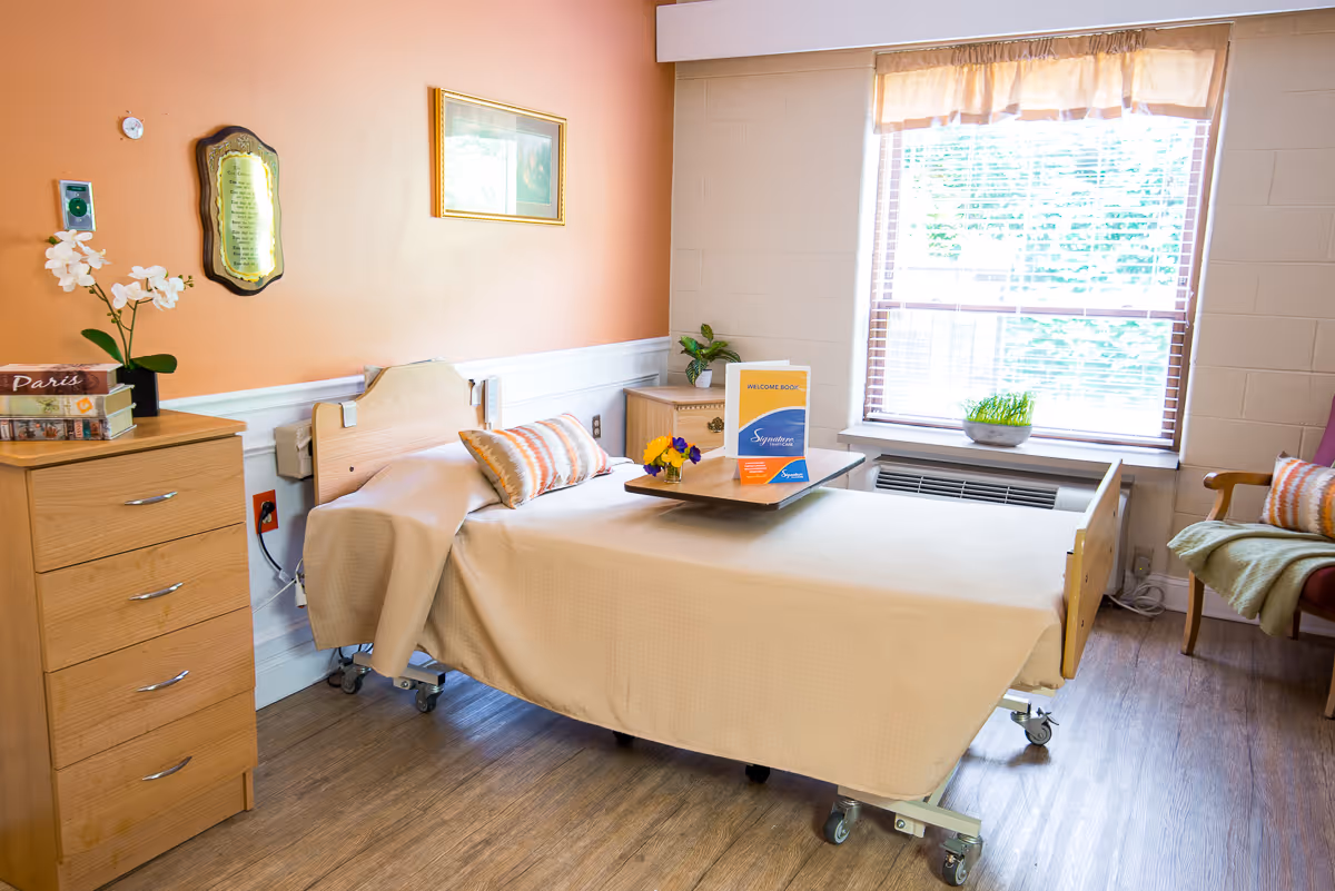 A bright and cozy bedroom in a senior living facility with a single bed covered in a beige blanket, a wooden dresser with books and a potted orchid, a small bedside table with a welcome book and flowers, a large window with blinds letting in natural light, and a comfortable chair with a cushion and blanket.