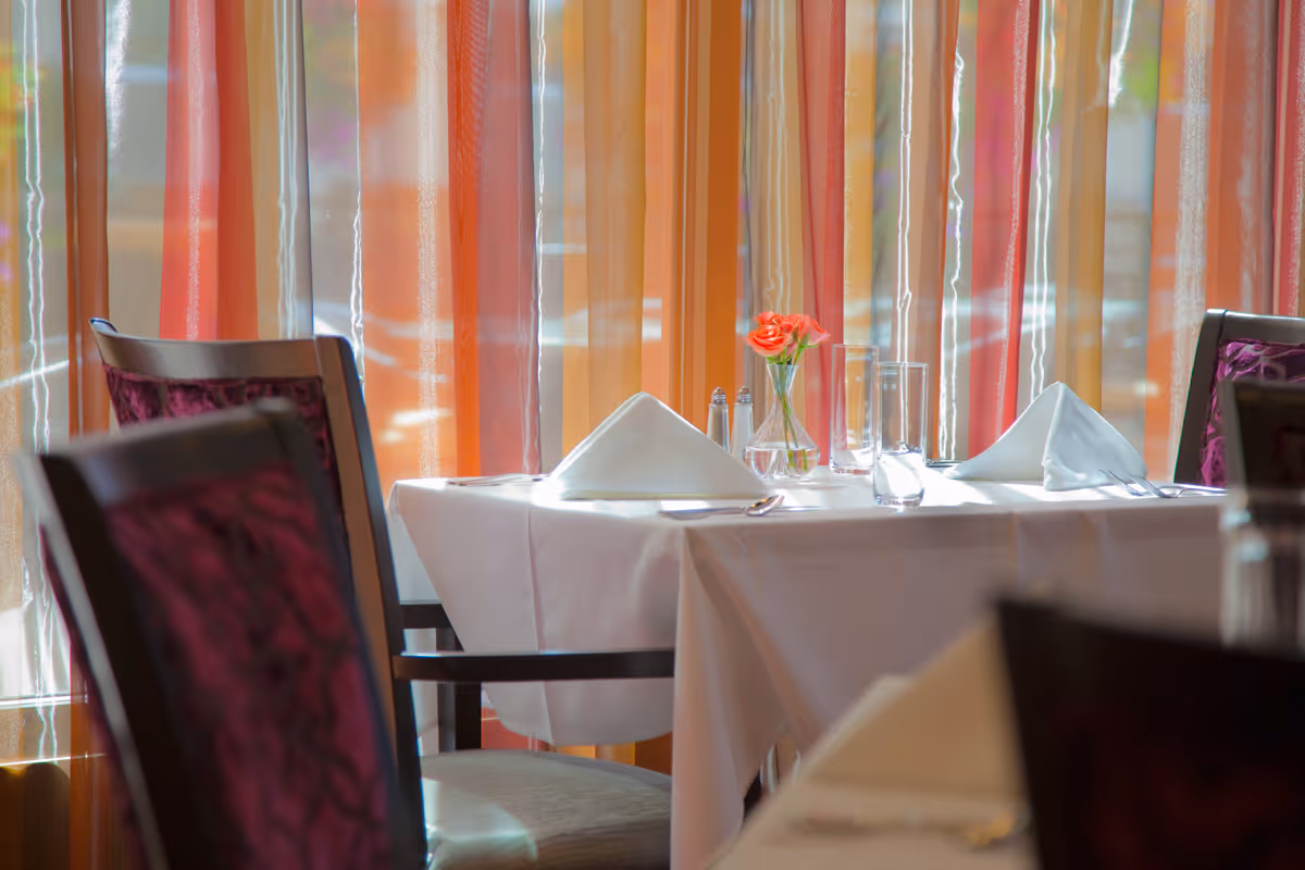 A dining table set for two with white tablecloth, folded white napkins, glassware, silverware, salt and pepper shakers, and a small vase with a red flower in front of colorful sheer curtains.