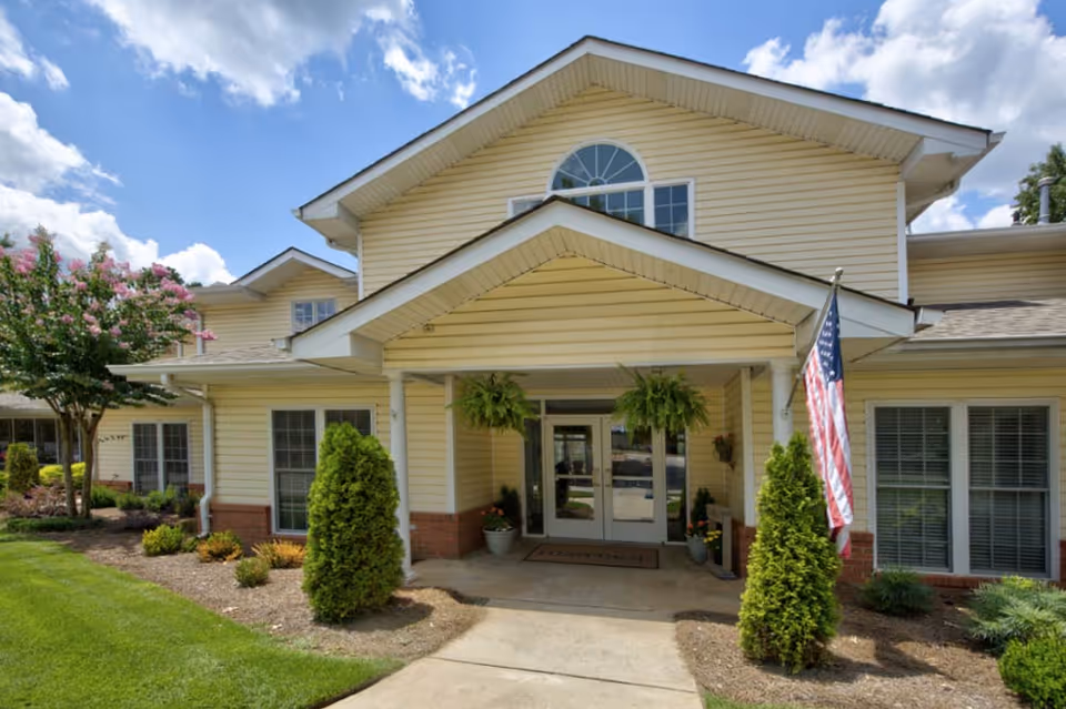 Front entrance of a yellow two-story senior living building with a covered portico, potted plants, and an American flag.