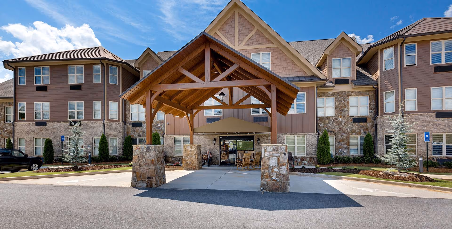 Front exterior view of Oaks at Acworth senior living facility with a covered entrance supported by stone pillars, a driveway, and a multi-story building with brown siding, stone accents, and multiple windows under a blue sky with clouds.