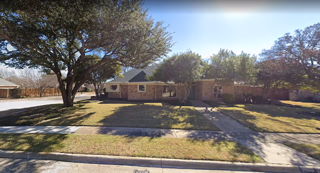 Single-story brick building with a sloped roof surrounded by a grassy lawn and several large trees, under a clear blue sky with sunlight filtering through the branches.