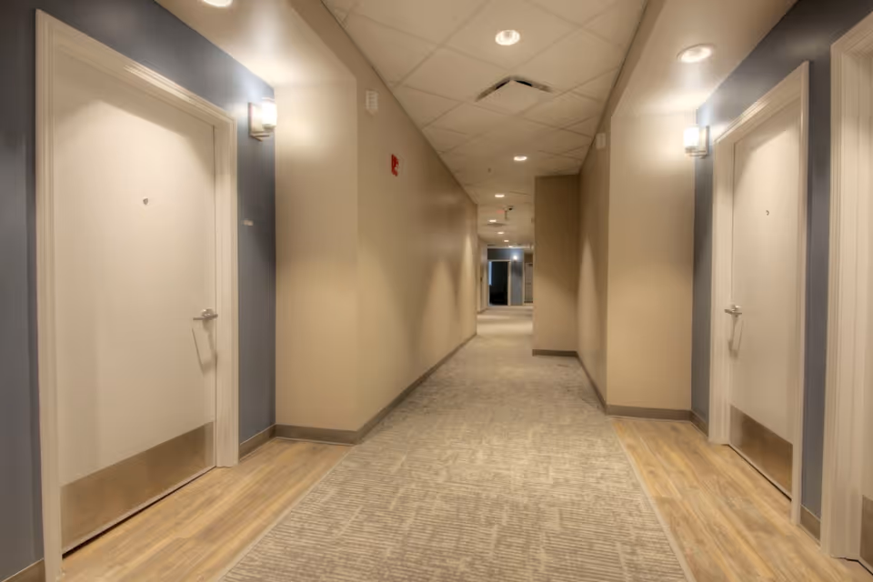 A well-lit hallway in Meadows Assisted Living and Care Campus with beige walls, carpeted floor, and two white doors on either side with metal kick plates. The ceiling has recessed lighting and a tiled pattern.
