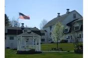 White multi-story building with a gazebo, American flag, trees, and a grassy lawn under a clear blue sky.