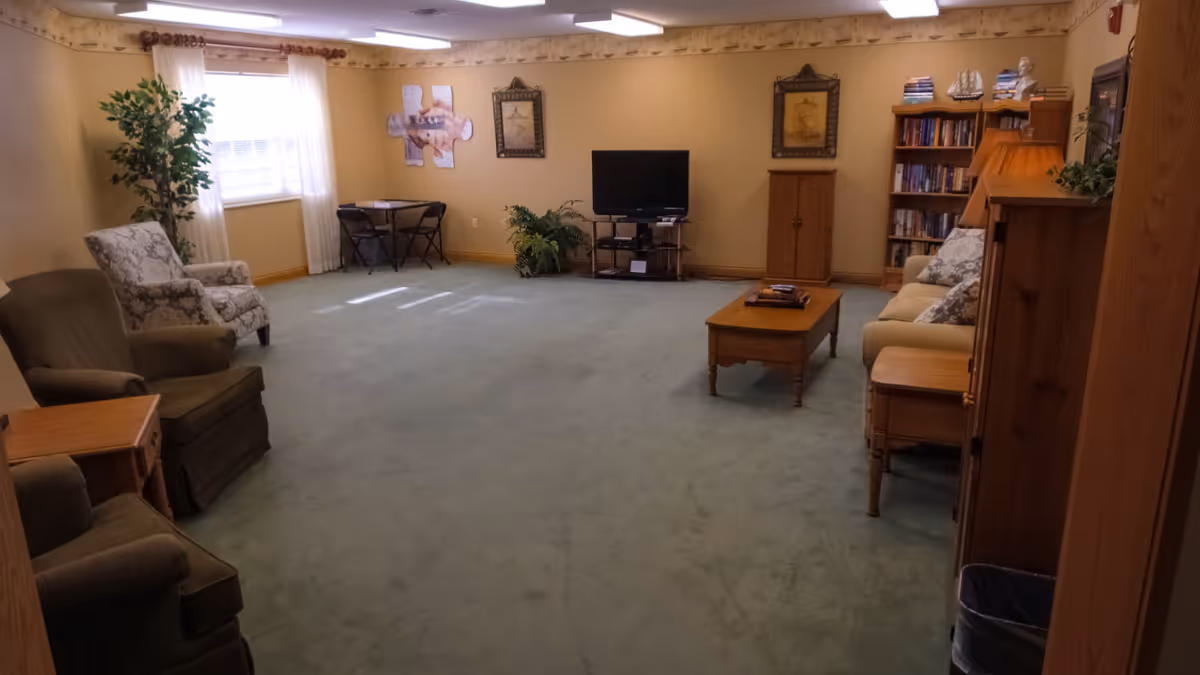 A spacious living room with green carpet, beige walls, and several pieces of furniture including armchairs, a sofa, a wooden coffee table, a TV on a stand, a bookshelf filled with books, and a small table with two chairs near a window with white curtains. There are framed pictures on the walls and some plants adding greenery to the room.