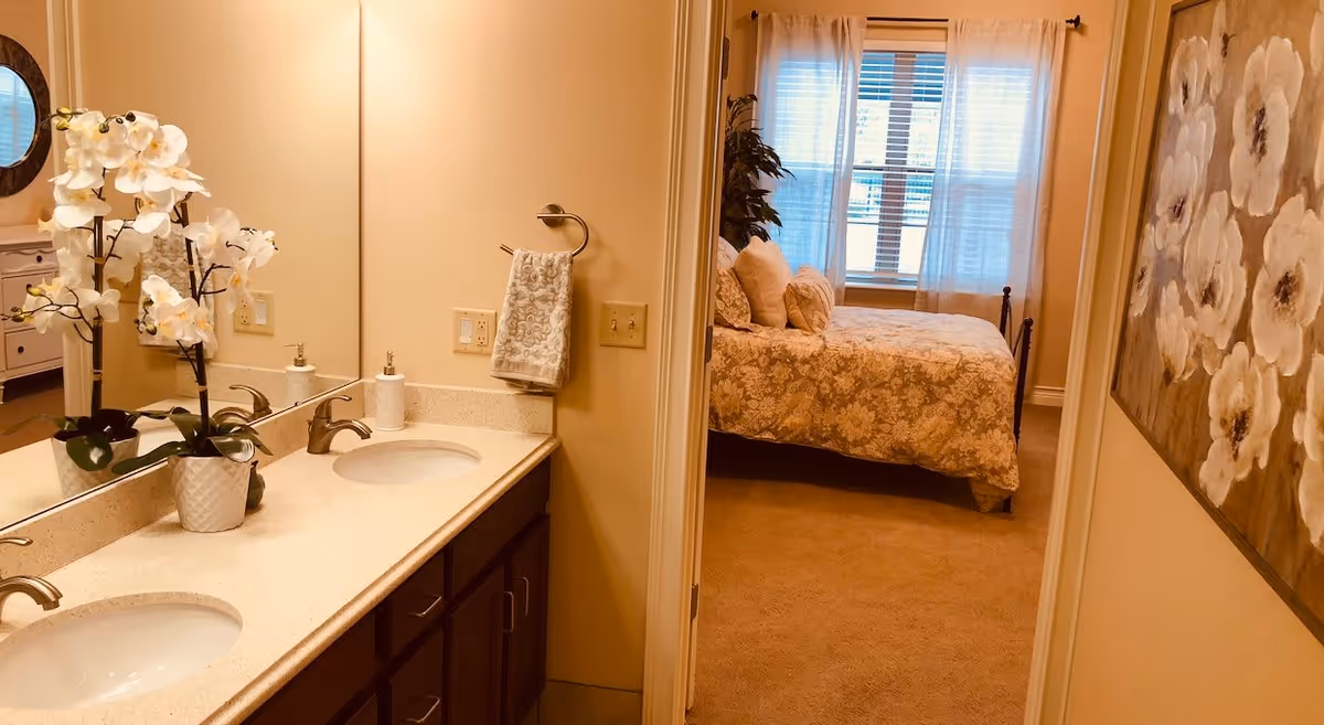 View of a bathroom with a double sink vanity, a large mirror, and a white orchid plant on the counter. Through an open doorway, a bedroom is visible with a bed covered in a floral bedspread, pillows, a window with sheer curtains, and a large floral painting on the wall.