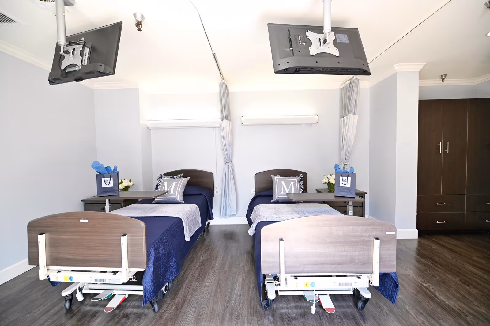 Two side-by-side hospital-style beds with navy bedding in a bright shared patient room with ceiling-mounted TVs and bedside tables.