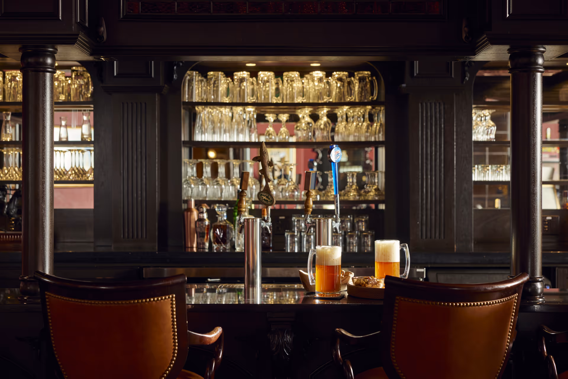 A dark wood bar with two leather bar stools in front. On the bar counter are two mugs of beer with foam, a bowl of snacks, and beer taps. Behind the bar are shelves filled with various glassware including mugs, wine glasses, and decanters, reflecting warm ambient lighting.