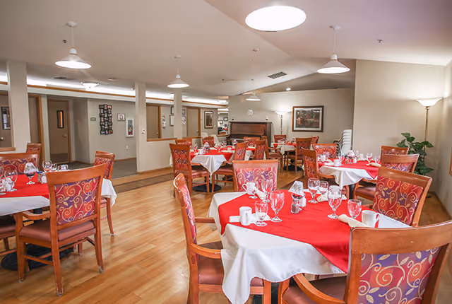 Dining room with round tables covered in red and white tablecloths, set with glassware and patterned wooden chairs on a hardwood floor.