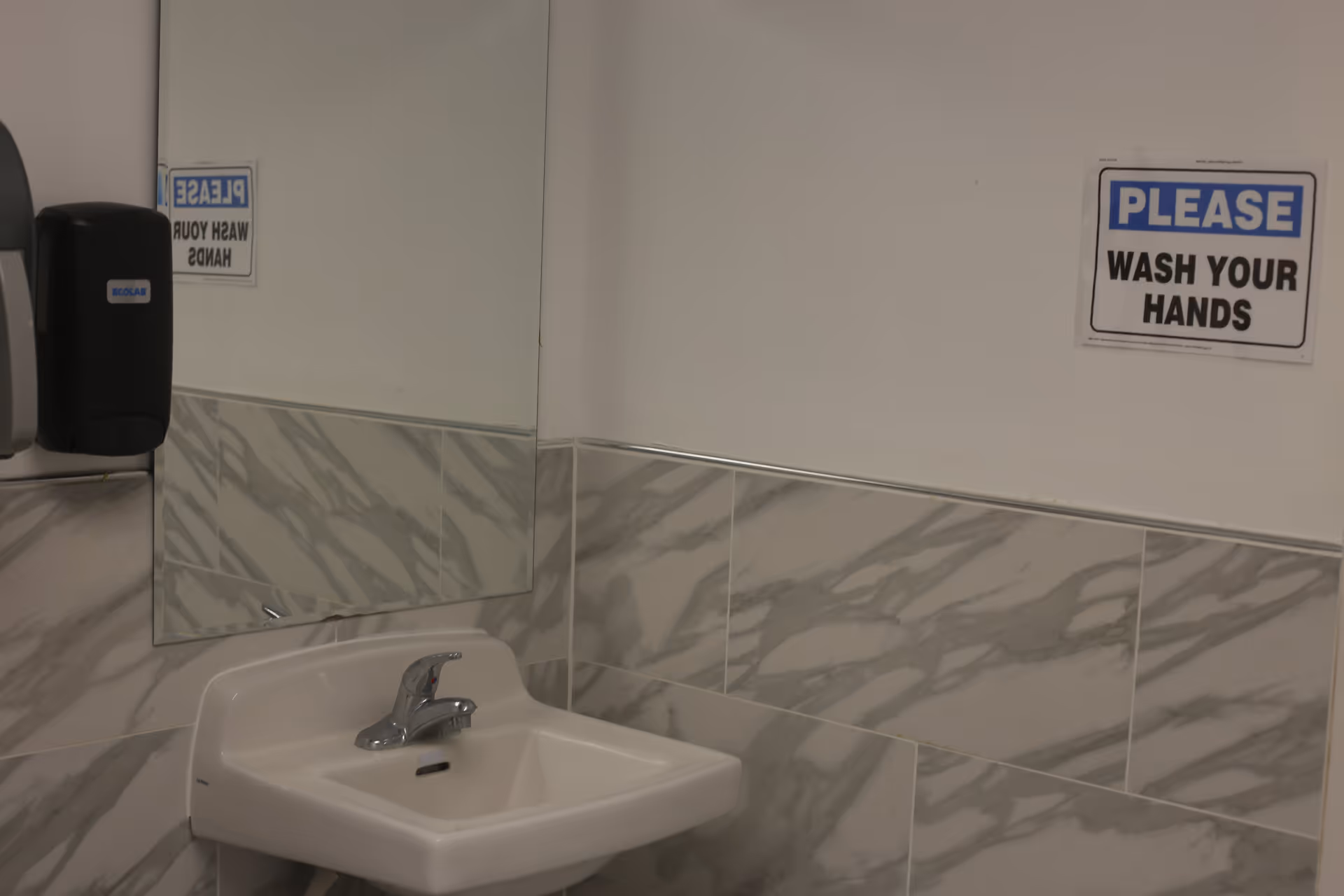 A small white sink with a silver faucet mounted on a wall with gray and white marble tiles. Above the sink is a rectangular mirror reflecting part of the sink and a black soap dispenser. On the white wall next to the sink is a sign that reads 'PLEASE WASH YOUR HANDS' in blue and black text.