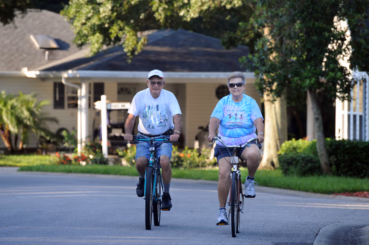 An elderly man and woman riding bicycles on a paved road in a residential area with houses and greenery in the background. The man is wearing a white cap, glasses, a white t-shirt, and blue shorts. The woman is wearing sunglasses, a blue tie-dye t-shirt, and dark shorts.
