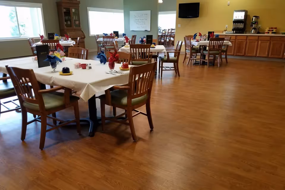 Dining room with several tables set with place settings and chairs, a beverage station and TV on the far wall.