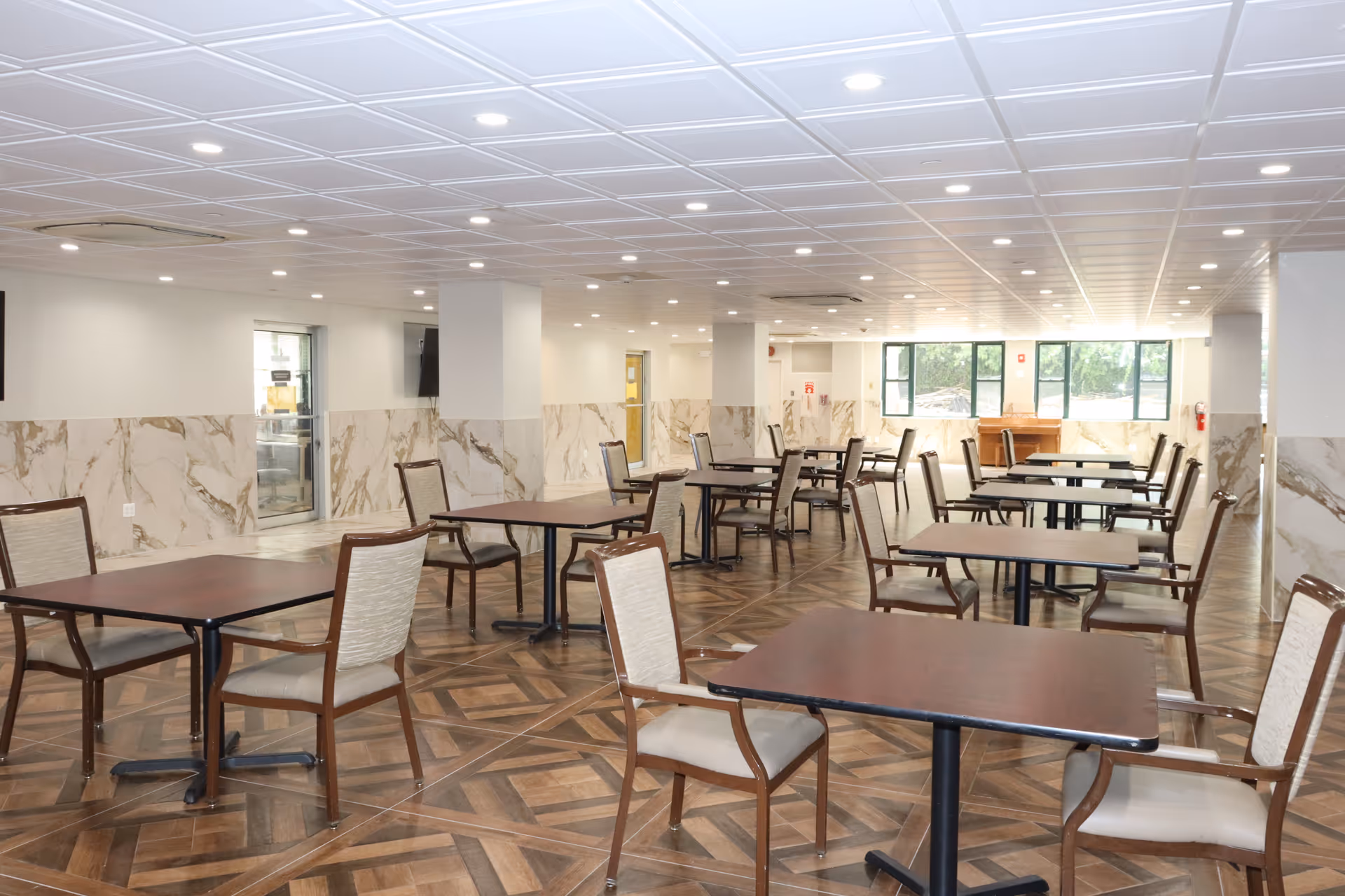 A spacious dining area with multiple square wooden tables and cushioned chairs arranged neatly. The room has a patterned wooden floor, white ceiling with recessed lighting, and walls partially covered with marble-like panels. Large windows at the far end allow natural light to enter the room.