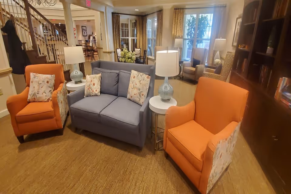 A cozy living room area in a senior living facility featuring a blue loveseat with floral pillows, two orange armchairs with floral sides, two white round side tables with blue lamps, a wooden bookshelf filled with books, and large windows with curtains letting in natural light.