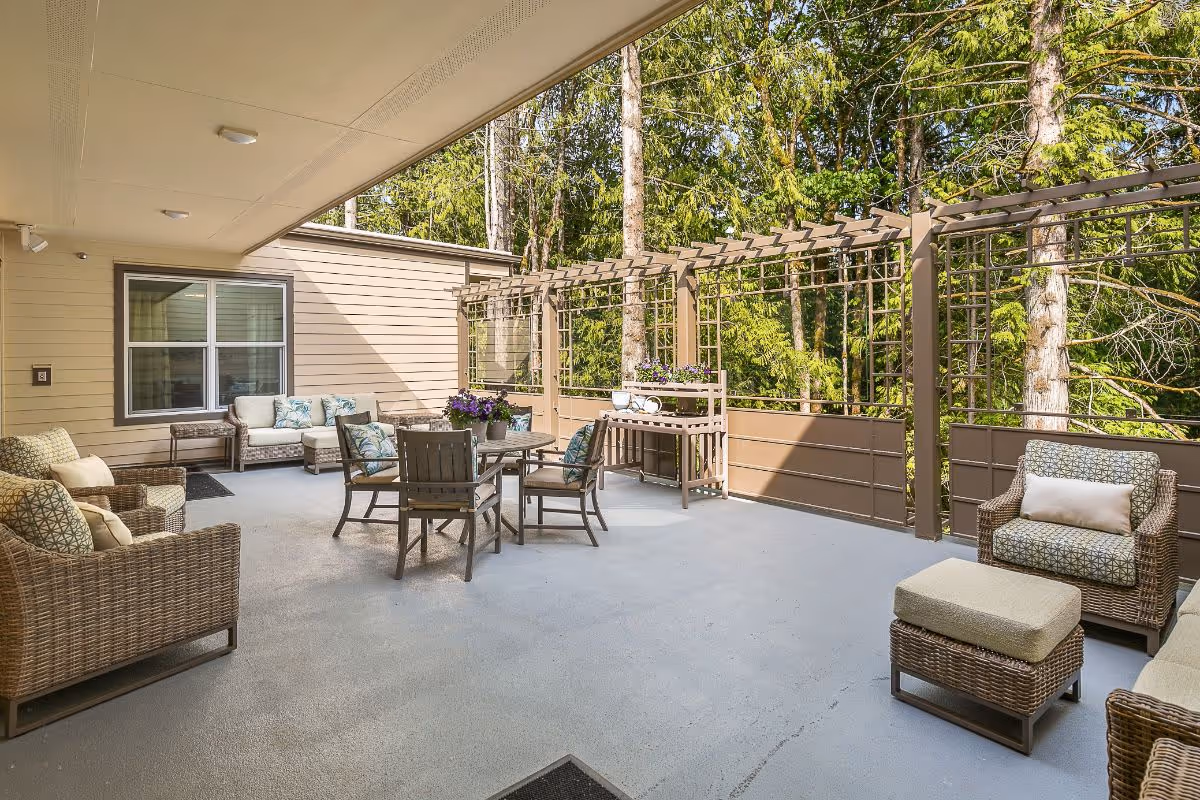 Outdoor patio area with wicker furniture including chairs, a loveseat, and a table with chairs. The patio is covered and has a trellis with potted plants and trees in the background.