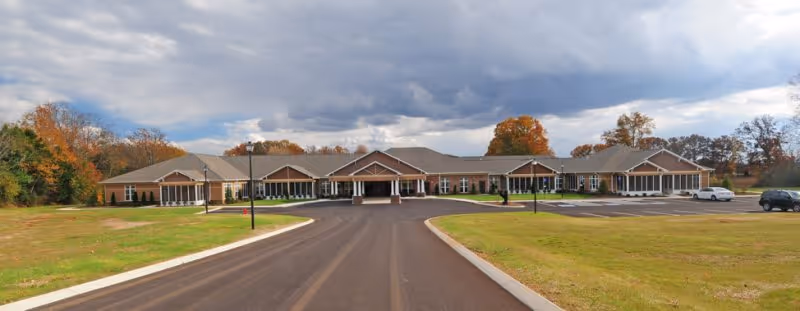 Wide front exterior view of Webb House Retirement Center, a single-story building with a large driveway and parking area, surrounded by grassy areas and trees with autumn foliage under a cloudy sky.
