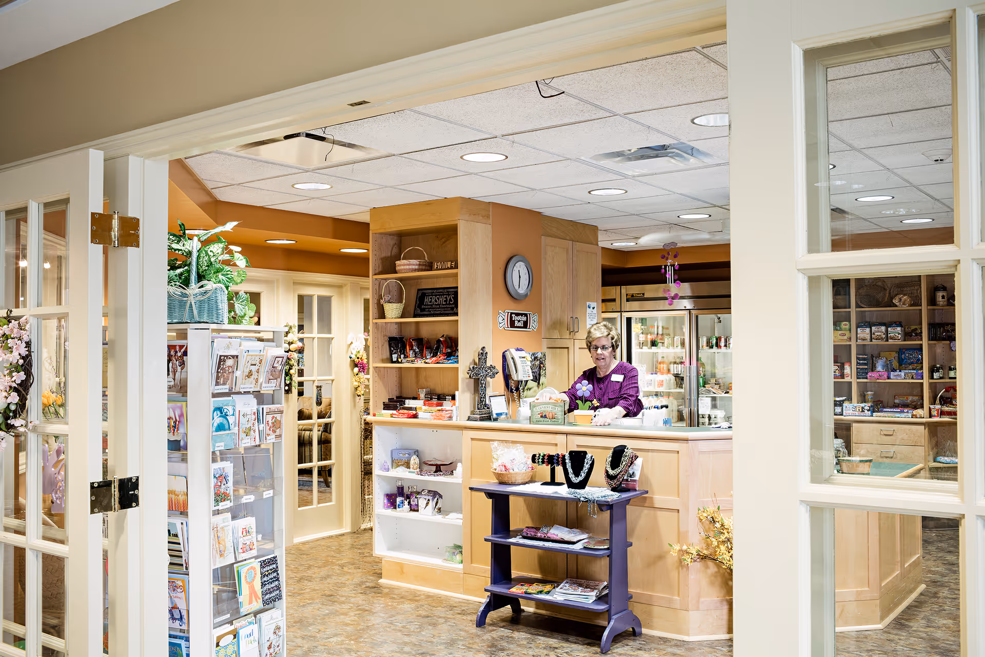 Interior view of a small shop or reception area inside a senior living facility. A woman stands behind a wooden counter with various items displayed, including jewelry on a small purple table in front. Shelves behind the counter hold snacks and other goods. To the left, a rotating rack displays greeting cards. The area is well-lit with ceiling lights and has a warm, welcoming atmosphere.