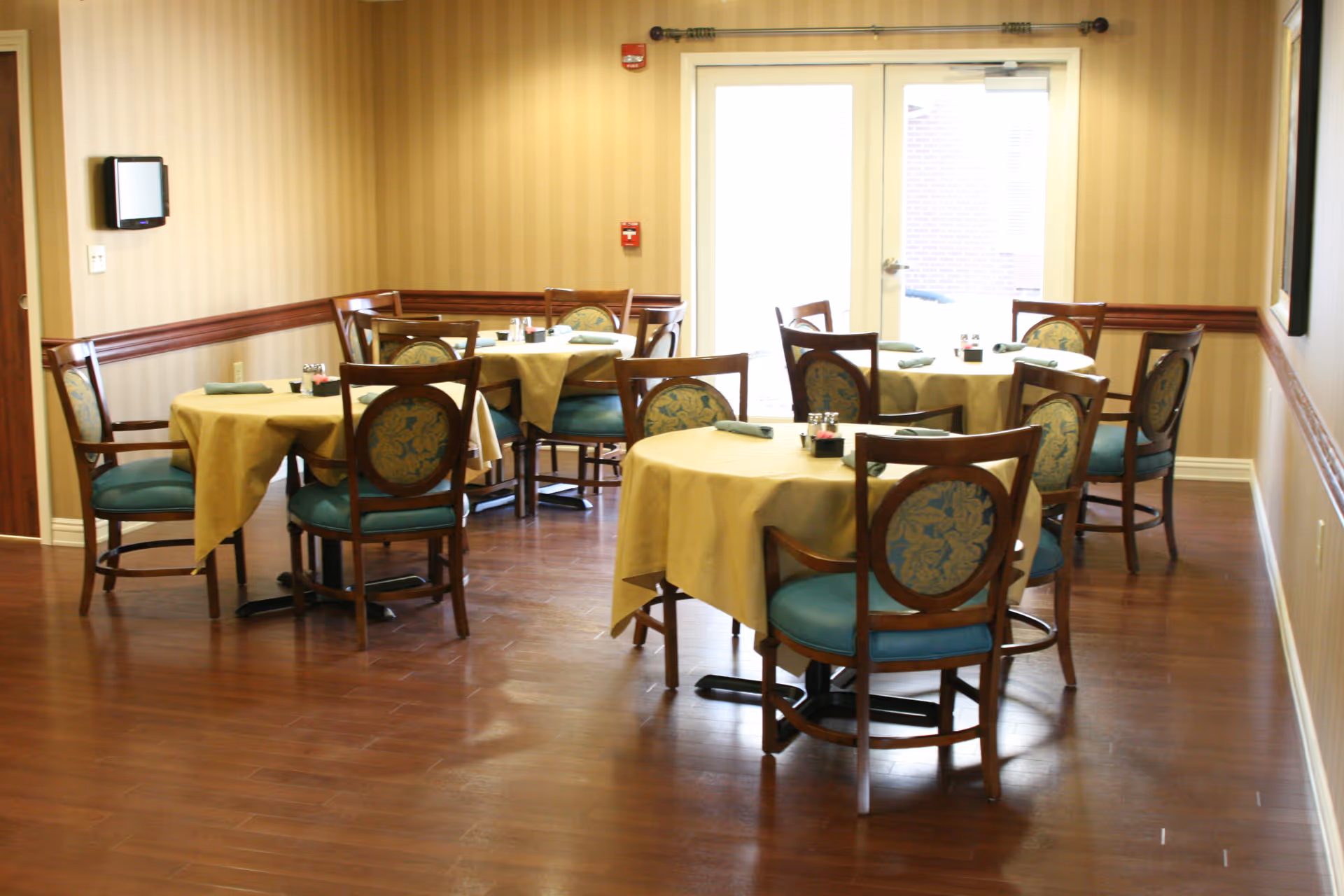 A dining room with four round tables covered with yellow tablecloths, each surrounded by wooden chairs with blue cushioned seats and floral patterned backs. The room has wooden flooring, beige striped wallpaper, and a glass door letting in natural light.