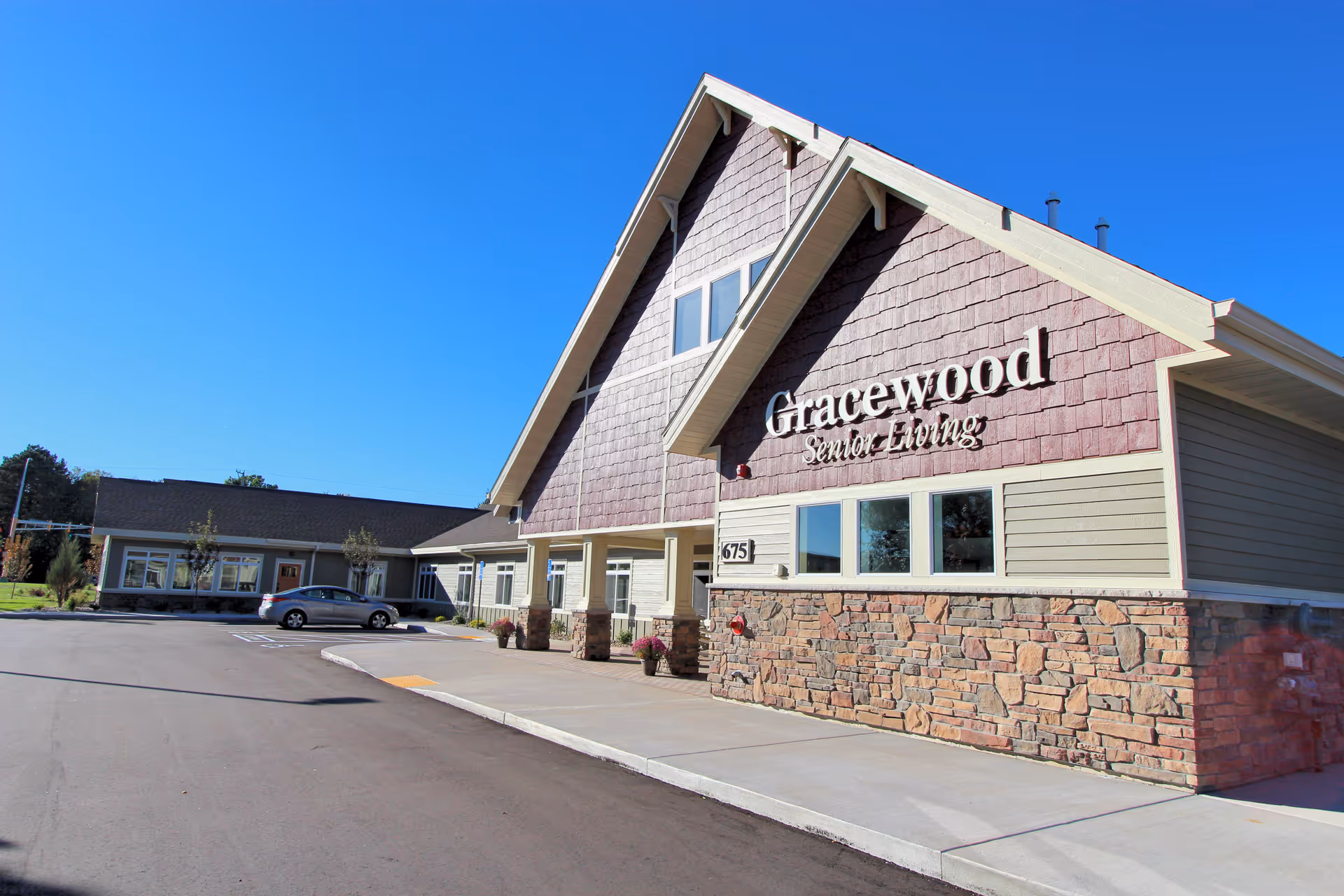 Exterior view of Gracewood Senior Living building with a stone and wood facade under a clear blue sky. A car is parked near the entrance and the building number 675 is visible.