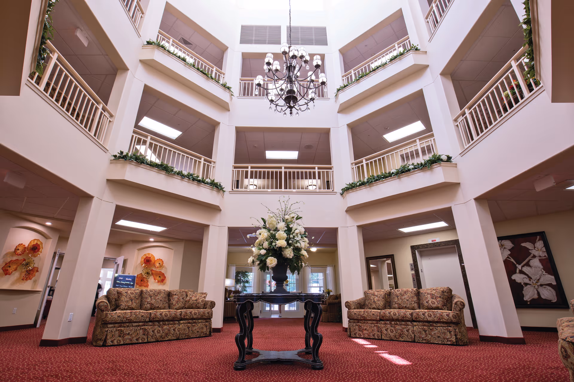 Spacious and well-lit interior common area of a senior living facility with a high ceiling and multiple balconies overlooking the space. The room features a central table with a large floral arrangement, two patterned sofas on either side, red carpet flooring, and decorative wall art. A chandelier hangs from the ceiling, adding elegance to the welcoming atmosphere.