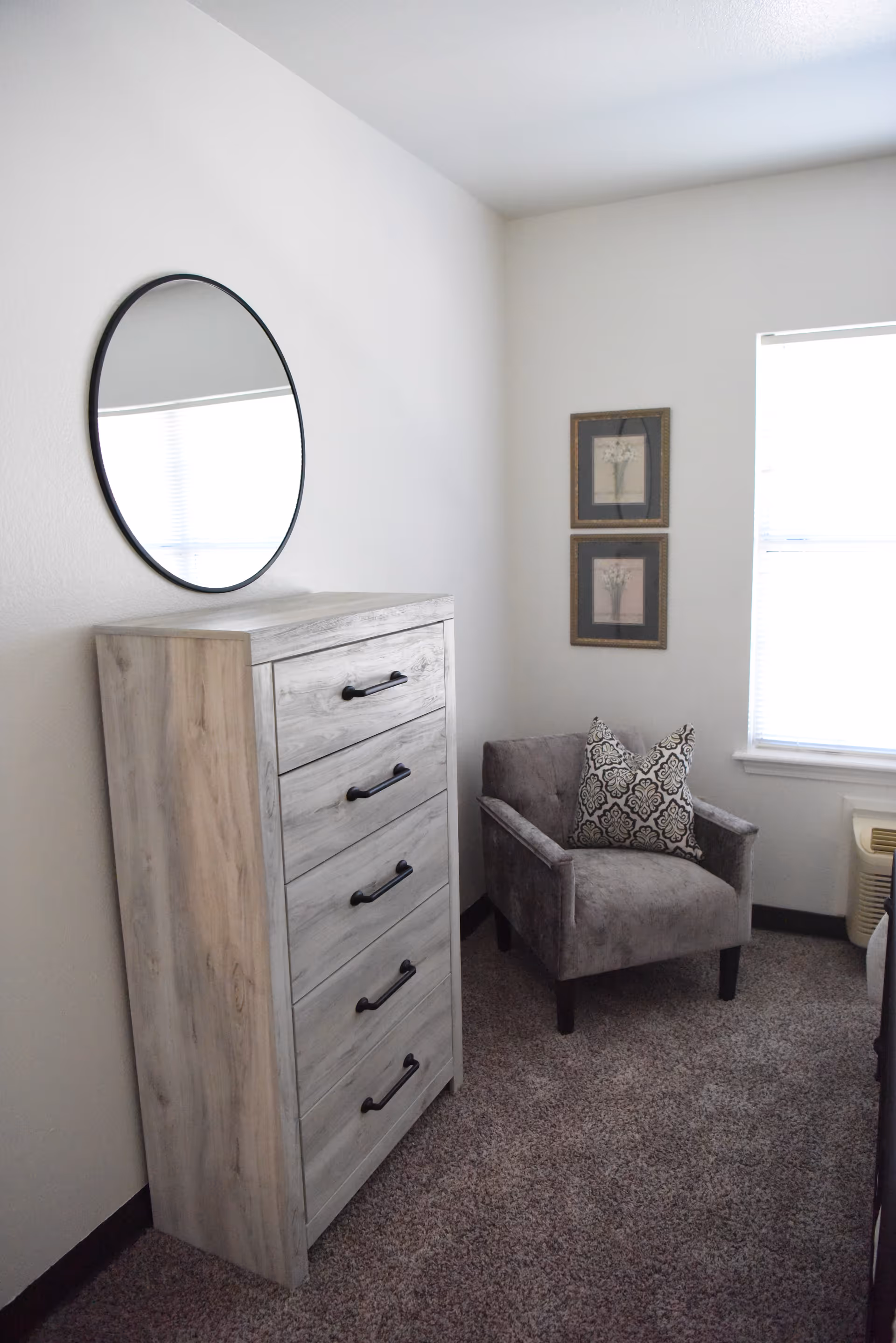 A corner of a bedroom featuring a tall wooden dresser with five drawers and black handles, a round mirror mounted on the wall above the dresser, a gray upholstered armchair with a decorative patterned pillow, two framed botanical prints on the wall, and a window with blinds letting in natural light.