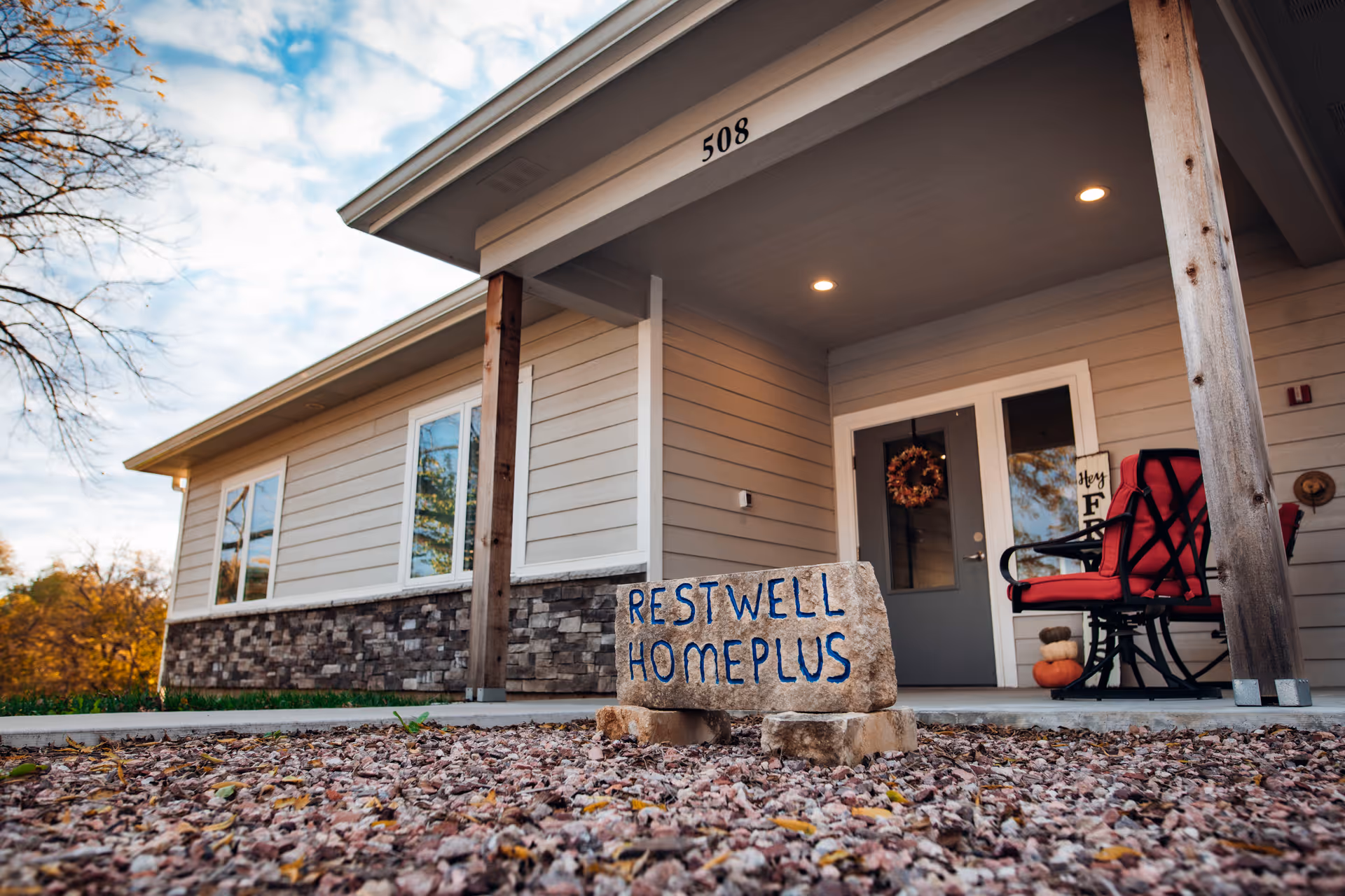 Exterior view of a single-story building with beige siding and stone accents. The entrance has a covered porch with two wooden posts, a red cushioned rocking chair, and a door decorated with a wreath. In front of the porch, there is a stone sign that reads 'RESTWELL HOMEPLUS'. The building number '508' is visible above the porch.