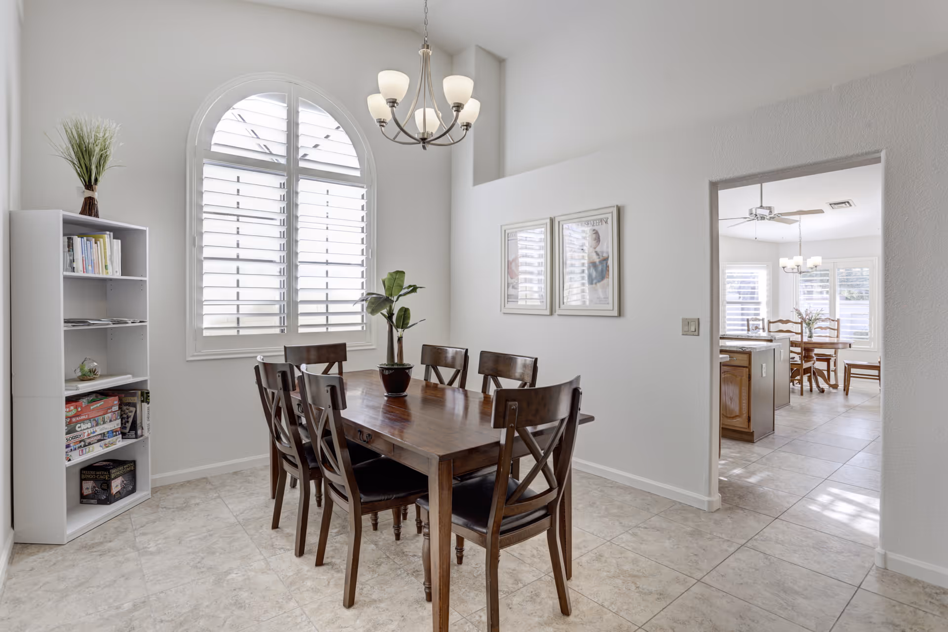 A bright dining room with a wooden table and six matching chairs. There is a large arched window with white shutters letting in natural light. A white bookshelf with books and board games stands in the corner, and two framed pictures hang on the wall. A chandelier with five lights hangs above the table. Through an open doorway, another dining area and kitchen are visible.