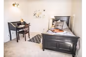 Small furnished bedroom with a single dark wood bed, desk and chair, lamp, and wall art.