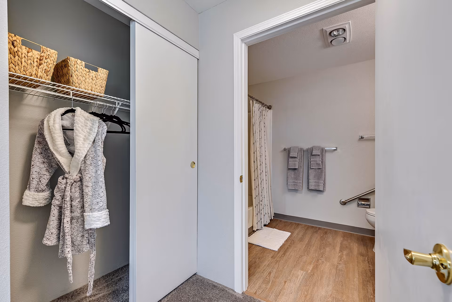 View of a closet with a hanging bathrobe and two woven baskets on a shelf, adjacent to a bathroom entrance. The bathroom features a shower with a curtain, two gray towels hanging on a towel rack, a grab bar near the toilet, and wood-style flooring.