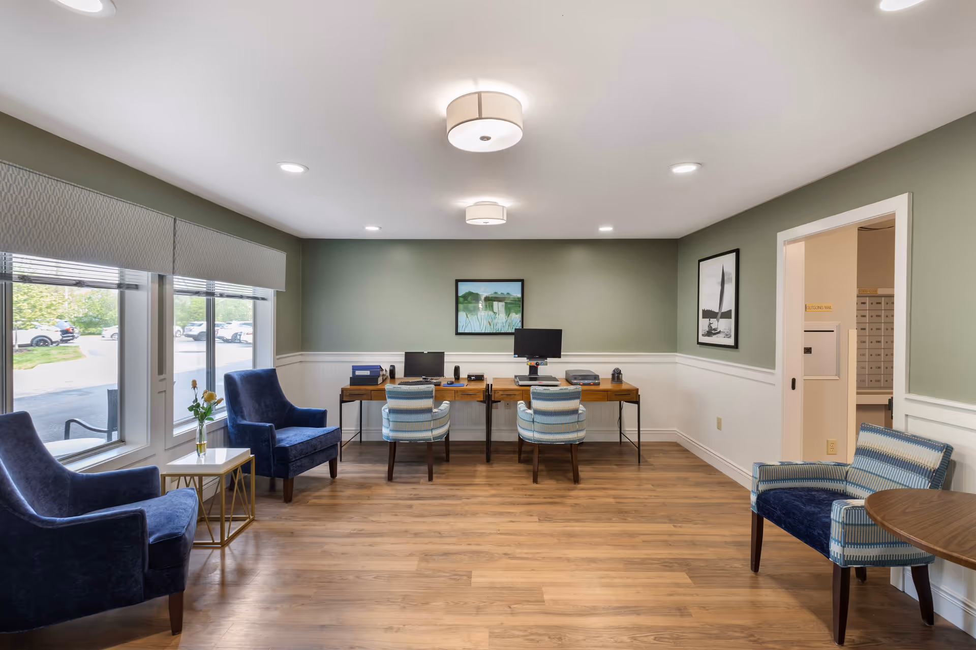 A well-lit office area in a senior living facility with two wooden desks against a green wall, each desk equipped with a computer and office supplies. There are two blue and white striped chairs in front of the desks and two blue armchairs near large windows on the left side. The floor is wooden, and there are framed pictures on the walls. A doorway on the right leads to a mailroom with mailboxes.