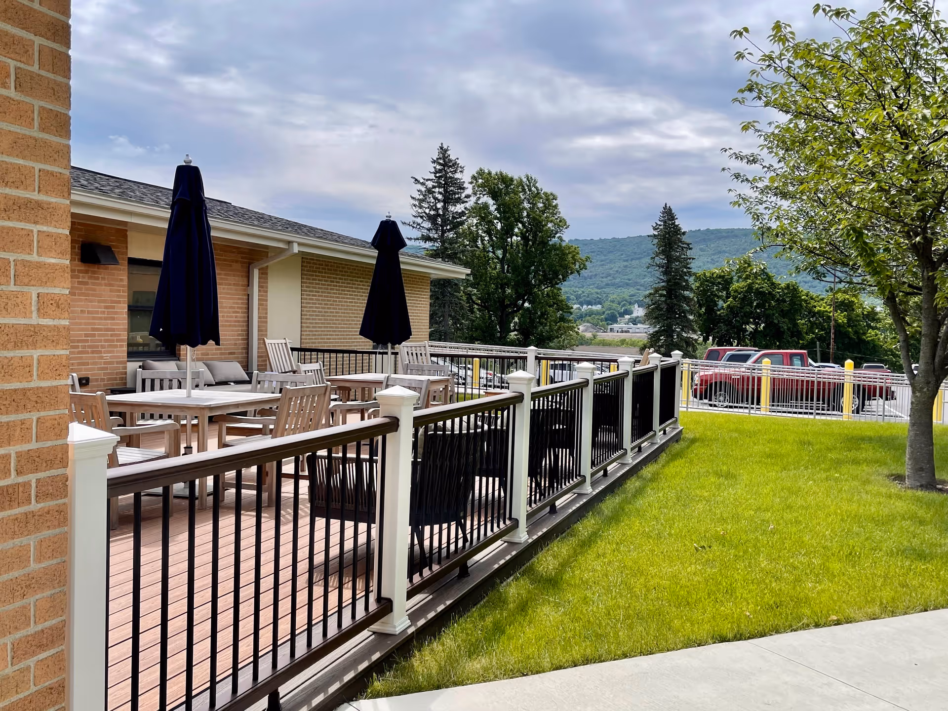 Outdoor patio area at Allied Services Meade Street Residence with wooden tables and chairs, two closed dark blue umbrellas, a black and white railing, green grass, a tree, and a view of distant hills under a cloudy sky.