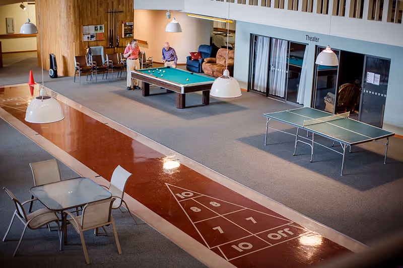 Indoor recreational area featuring a shuffleboard court, a pool table with two elderly people playing, a ping pong table, and a seating area with a glass table and chairs. The area also has a theater room with glass doors and comfortable seating nearby.