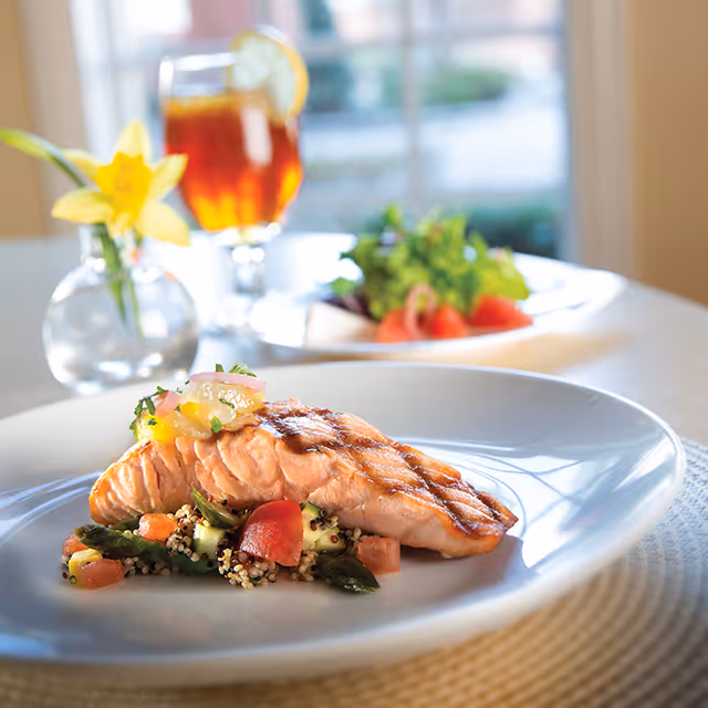 A plated grilled salmon fillet served on a bed of quinoa salad with vegetables, accompanied by a glass of iced tea with a lemon slice and a small vase with a yellow flower on a table near a window.