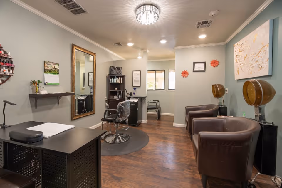 Interior view of a salon area in a senior living facility with a manicure table, a large wall mirror, salon chairs, hair dryers, and shelves with hair care products. The room has wooden flooring, light green walls, and decorative wall art.