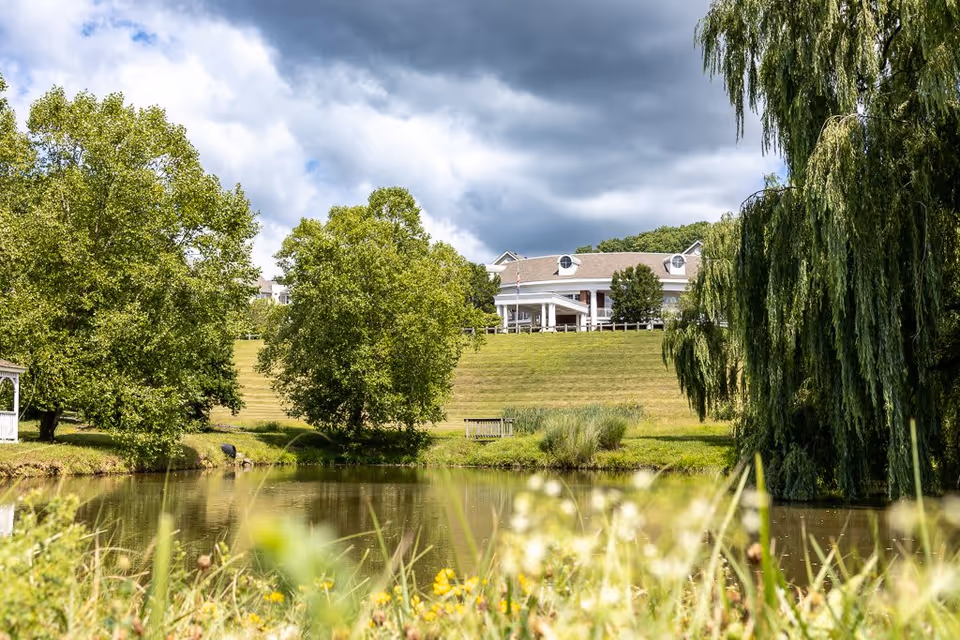 A scenic view of a pond surrounded by green trees and grass with a large building in the background under a cloudy sky. The building is situated on a hill with a flagpole and a bench near the pond.