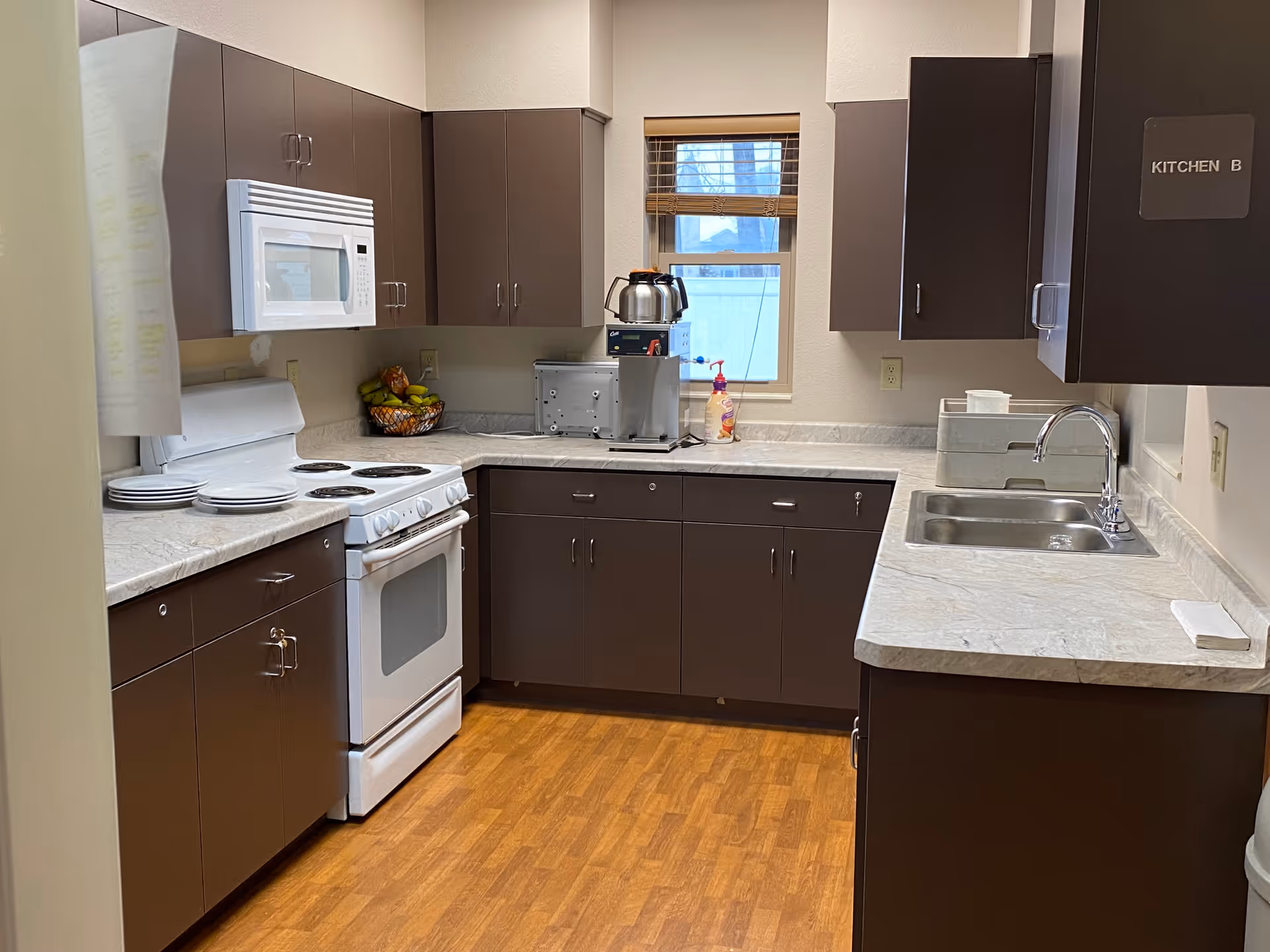 A clean kitchen with dark brown cabinets, a white stove with oven, a white microwave above the stove, a double sink, and a coffee maker on the countertop. There is a small window with blinds above the counter, and a basket of fruit on the counter. The floor is wooden, and a sign on one cabinet reads 'KITCHEN B'.