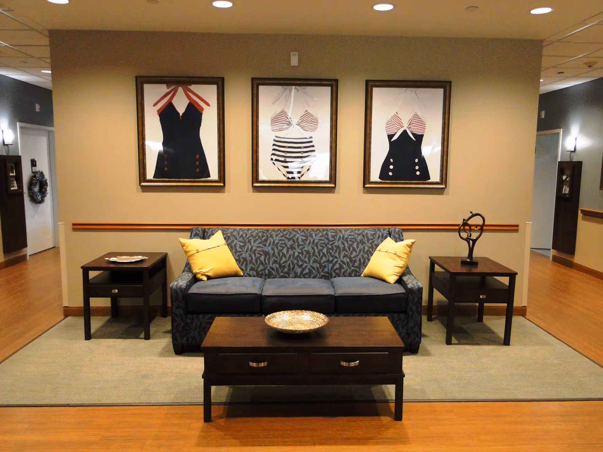 A cozy seating area in an assisted living facility featuring a patterned blue sofa with two yellow pillows, flanked by two dark wooden side tables. A dark wooden coffee table with a decorative bowl is placed in front of the sofa. Above the sofa, three framed pictures of vintage swimsuits are hung on a beige wall. The floor is wooden with a large area rug under the furniture, and the space is softly lit by ceiling lights.