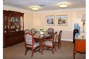 A dining room with a wooden dining table surrounded by six upholstered chairs with floral patterns. A wooden china cabinet is against the left wall, and two framed landscape paintings hang on the beige wall. There is a water cooler and a lamp on a wooden side table on the right side of the room. The room is carpeted and has ceiling lights.