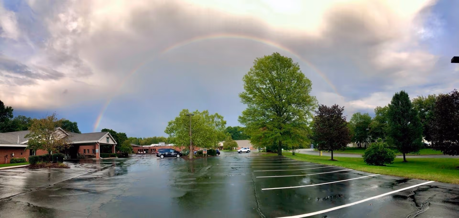 A wet parking lot in front of a brick building with a covered entrance, surrounded by green trees and grass. A rainbow arches across a cloudy sky above the scene.