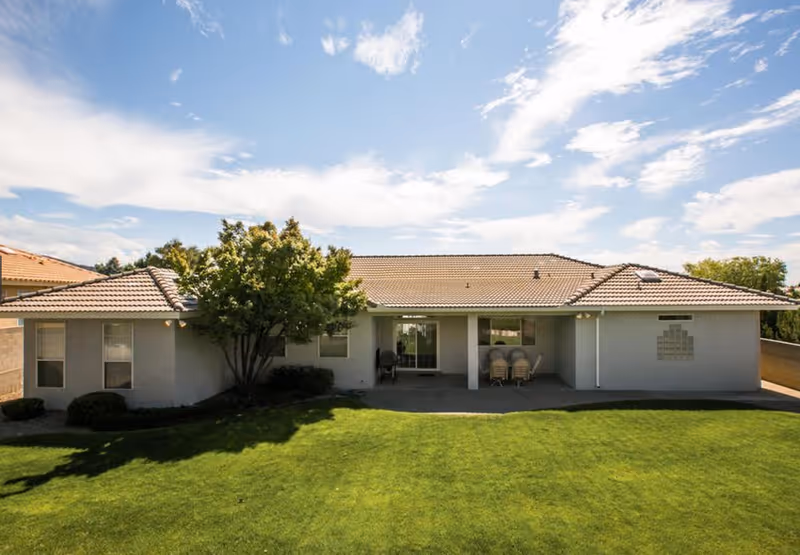 Single-story assisted living building with a tiled roof, covered patio, tree and a well-kept green lawn under a blue sky.