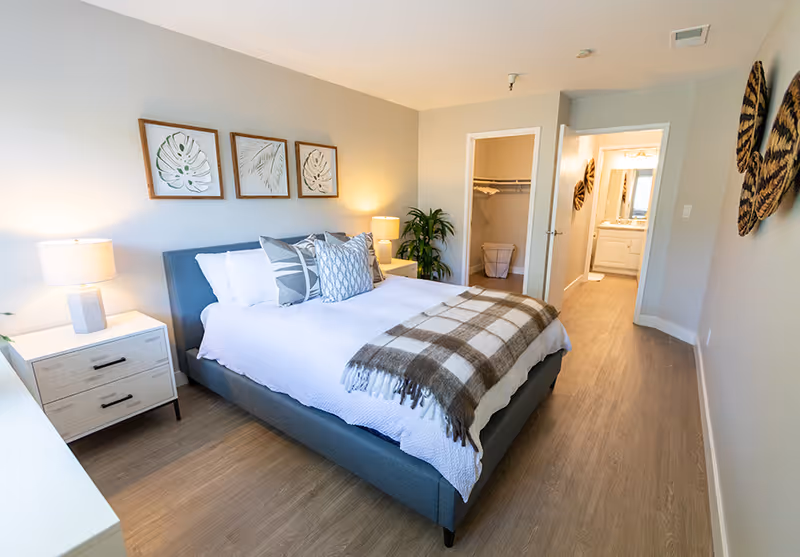 A bright and modern bedroom with a large bed featuring white bedding and multiple decorative pillows. There is a plaid throw blanket at the foot of the bed. Two bedside tables with lamps flank the bed. Three framed botanical prints hang above the bed. The room has light wood flooring and neutral-colored walls. An open door reveals a walk-in closet with a laundry basket, and another door leads to a bathroom with a vanity and mirror. Decorative wall art is visible on the right wall.