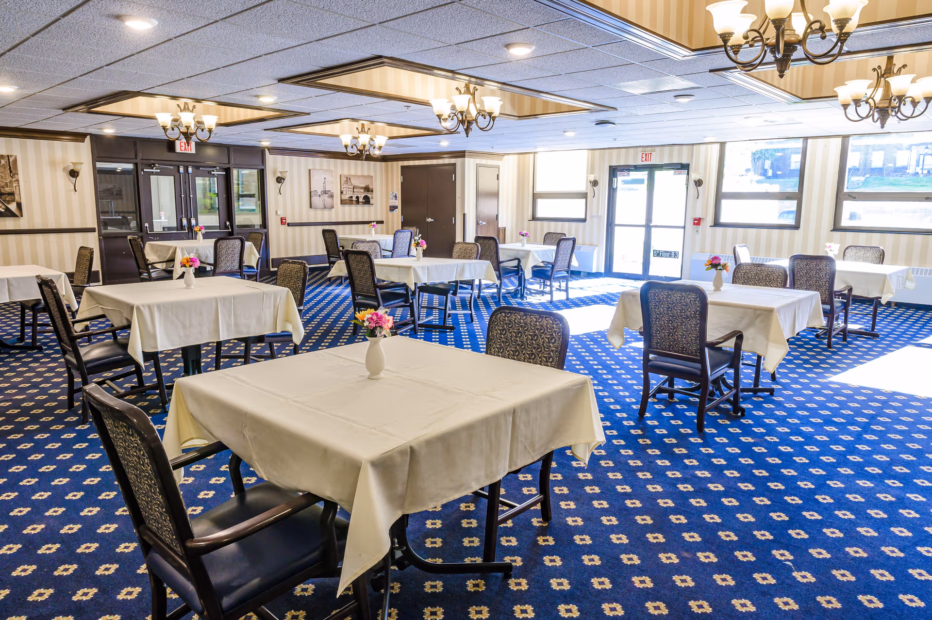 A bright dining room with multiple tables covered in white tablecloths, each adorned with a small vase of flowers. The room features patterned blue carpet, several chandeliers hanging from the ceiling, and large windows letting in natural light. There are framed pictures on the walls and double doors at the far end of the room.