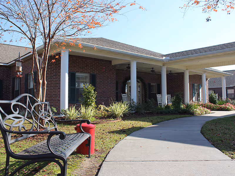 Brick building front with a covered columned porch, rocking chairs, a curved sidewalk, bench, and landscaping.