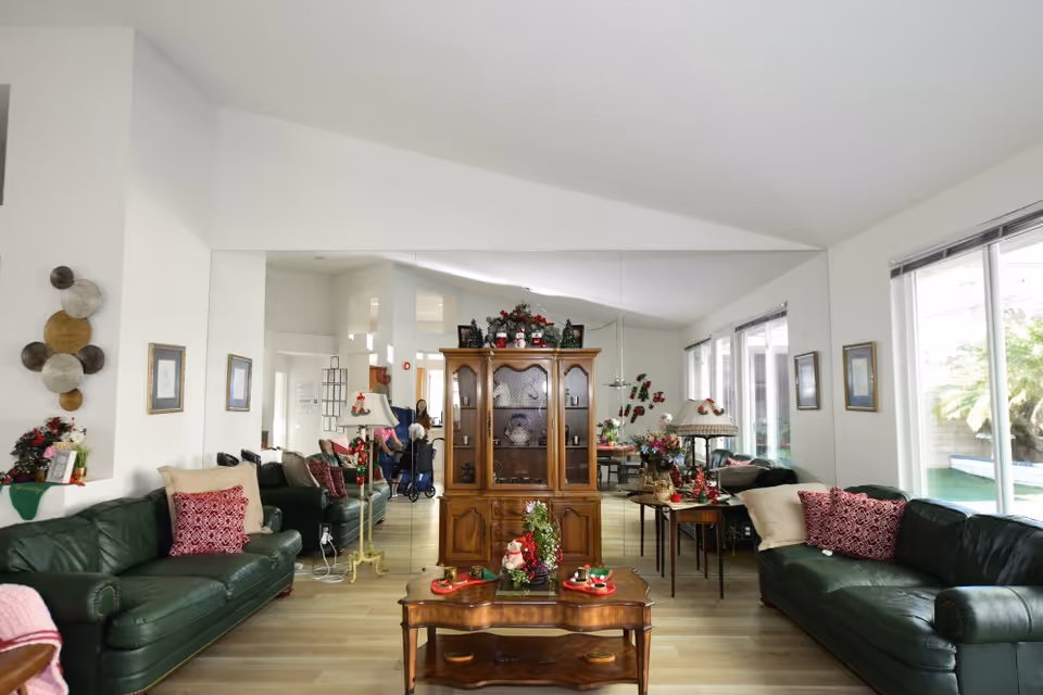 A bright living room with two dark green leather sofas adorned with red and beige pillows. A wooden coffee table with holiday decorations is in the center. Behind the table is a wooden cabinet with glass doors, also decorated for the holidays. The room has large windows on the right side letting in natural light and a large mirror on the back wall reflecting the room. There are framed pictures on the walls and a floor lamp beside one of the sofas. A person in a wheelchair is visible in the background near the entrance.