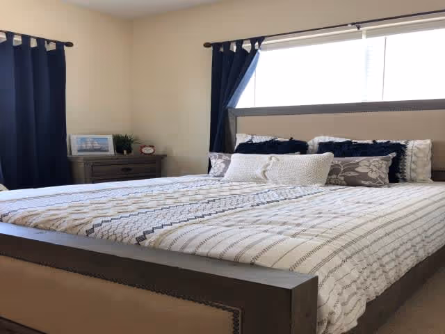 A neatly made bed with a patterned white and gray bedspread, several decorative pillows, and a wooden headboard. There is a window with dark blue curtains behind the bed and a wooden nightstand with a small plant, a framed picture, and a clock on it.