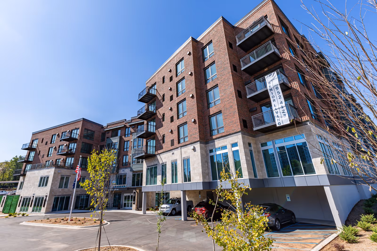 Exterior view of a modern multi-story senior living facility with brick and stone facade, balconies, and large windows under a clear blue sky. There are a few young trees and parked cars in front of the building.