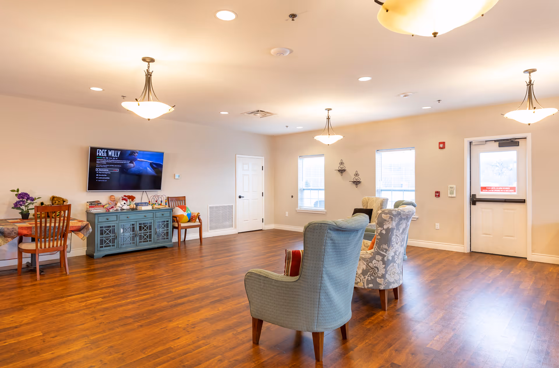 A bright and spacious common area in an assisted living facility with wooden flooring, several upholstered armchairs arranged in a semi-circle, a small table with chairs and toys, a wall-mounted TV displaying a movie titled 'Free Willy', and large windows letting in natural light.