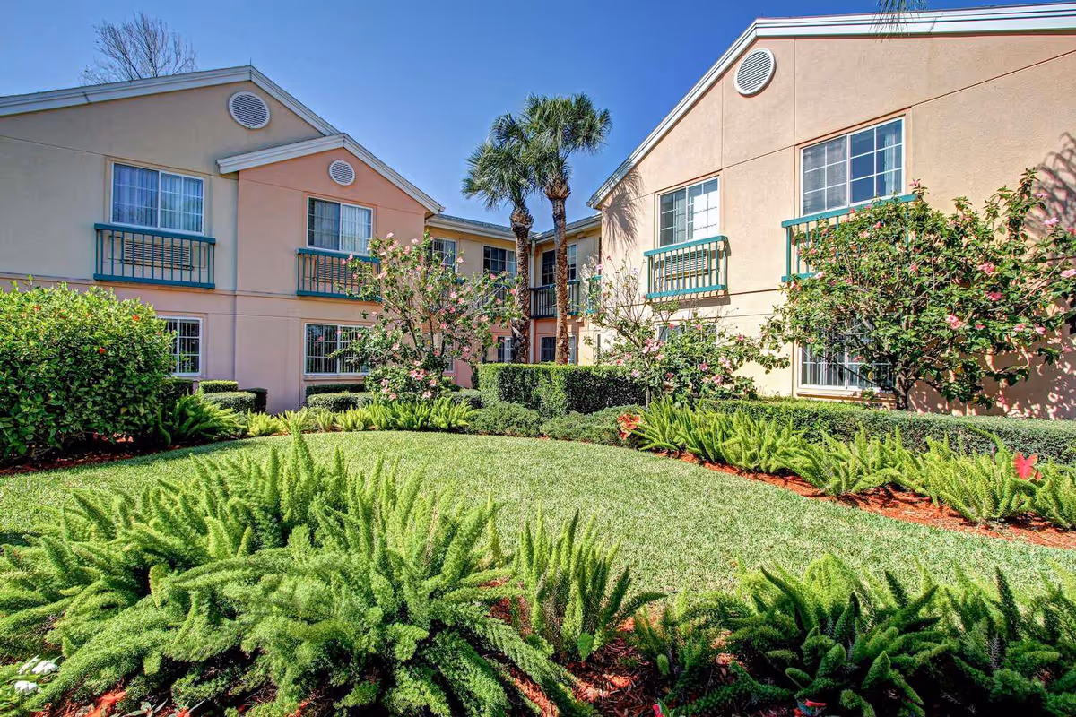 Two-story beige senior living building with small balconies around a manicured courtyard of palm trees, shrubs, and green lawn.